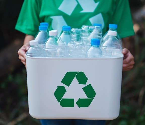 A woman collecting garbage and holding a recycle bin with plastic bottles in the outdoors